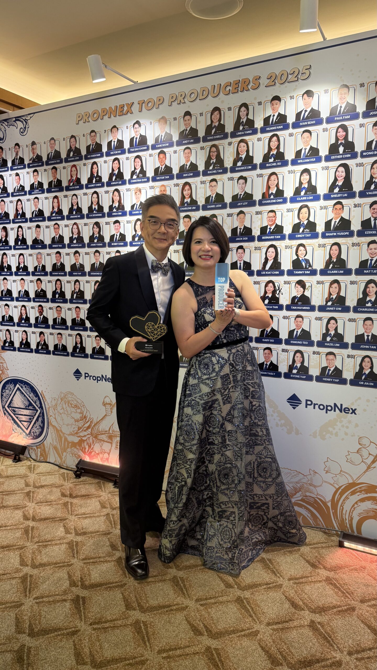 Two attendees holding awards in front of the PropNex Top Producers 2025 display.